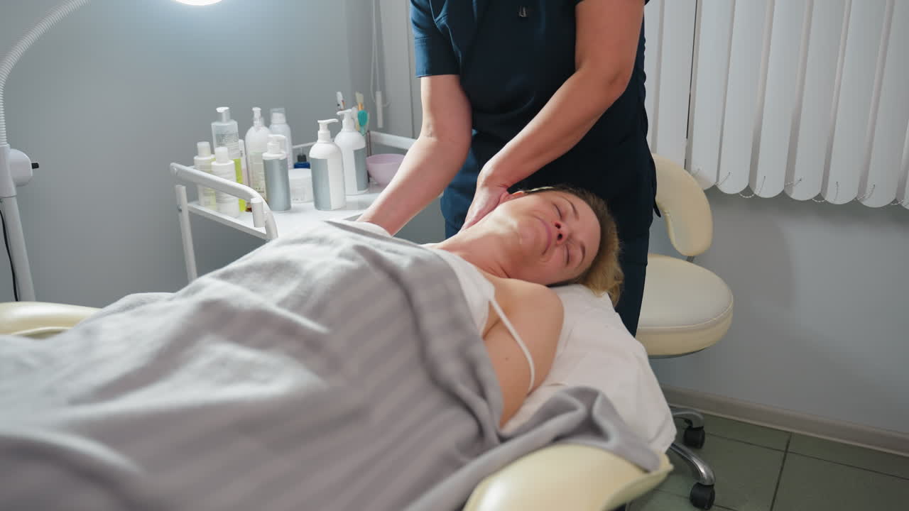 Wellness technician beside fair skinned lady gliding fingertips along side of patient neck toward shoulder during relaxing spa session in bright clinic room with subtle soft product trolley