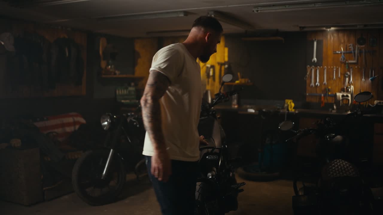 A blond guy in a white T-shirt inspects his motorcycle in a workshop garage