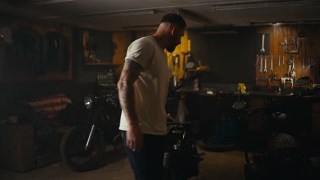 A-blond-guy-in-a-white-T-shirt-inspects-his-motorcycle-in-a-workshop-garage