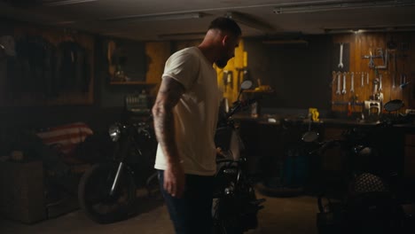 a blond guy in a white t-shirt inspects his motorcycle in a workshop garage