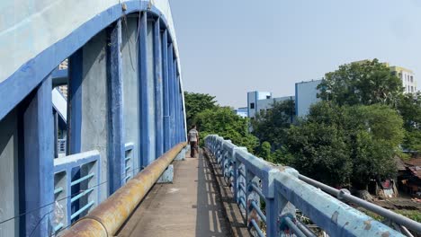 a poor man walking alone in a old blue color british bridge in kolkata with building and trees in background