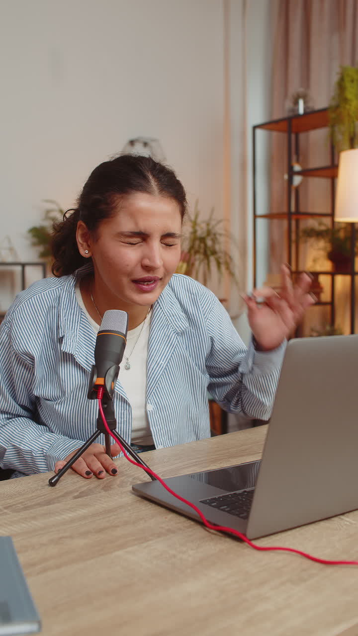 Joven influencer india feliz hablando por micrófono y webcam de portátil en la mesa de su oficina en casa