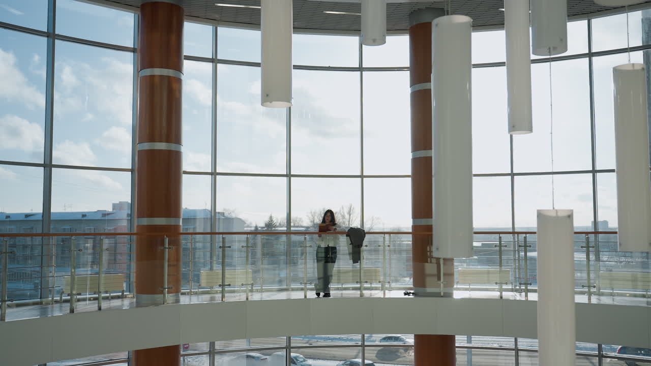 Full view of lady leaning casually on railing inside sunlit modern mall with glass windows, jacket resting beside her, urban background with cars moving along road outside enhances city atmosphere