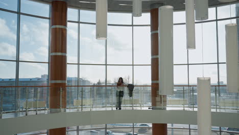 full view of lady leaning casually on railing inside sunlit modern mall with glass windows, jacket resting beside her, urban background with cars moving along road outside enhances city atmosphere