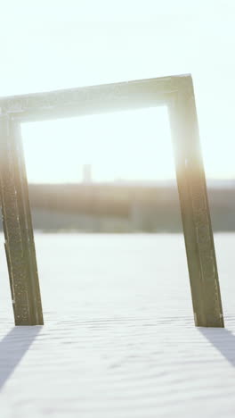 a weathered wooden frame stands alone in the sand, with the sun setting in the background.