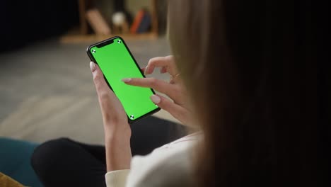 mujer usando un teléfono inteligente con pantalla verde. persona femenina irreconocible deslizando en el teléfono móvil con llave chroma, sentada en el sofá en la sala de estar. compras en línea, navegación por internet. tiempo libre en casa