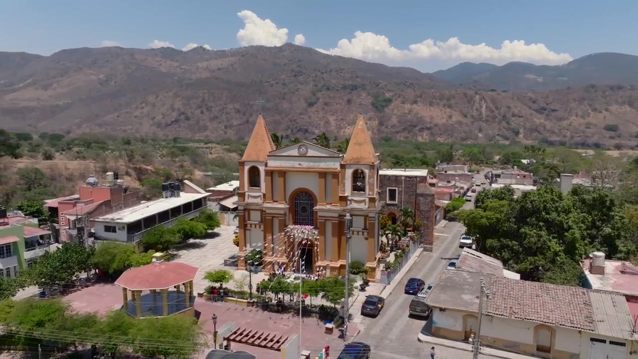 Kretsande flygfoto över Platanars centrum, med utsikt över kyrkfasaden till Parroquia de la Virgen de Platanar, det centrala torget och de omgivande bergen i Jalisco, Mexiko, en solig dag.
