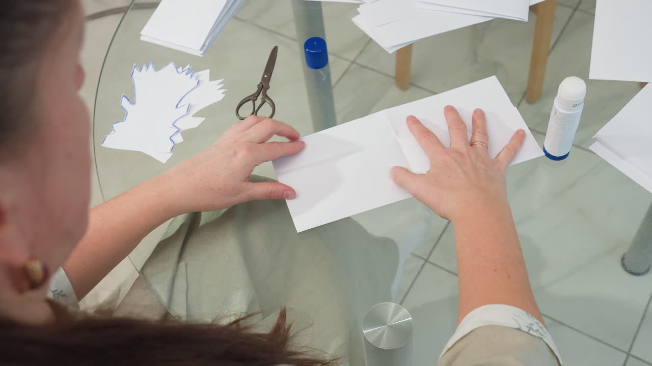 Back view of woman picking up paper from table, applying glue stick to it, preparing it for crafting, with scissors and other paper pieces in the background