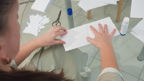 back view of woman picking up paper from table, applying glue stick to it, preparing it for crafting, with scissors and other paper pieces in the background