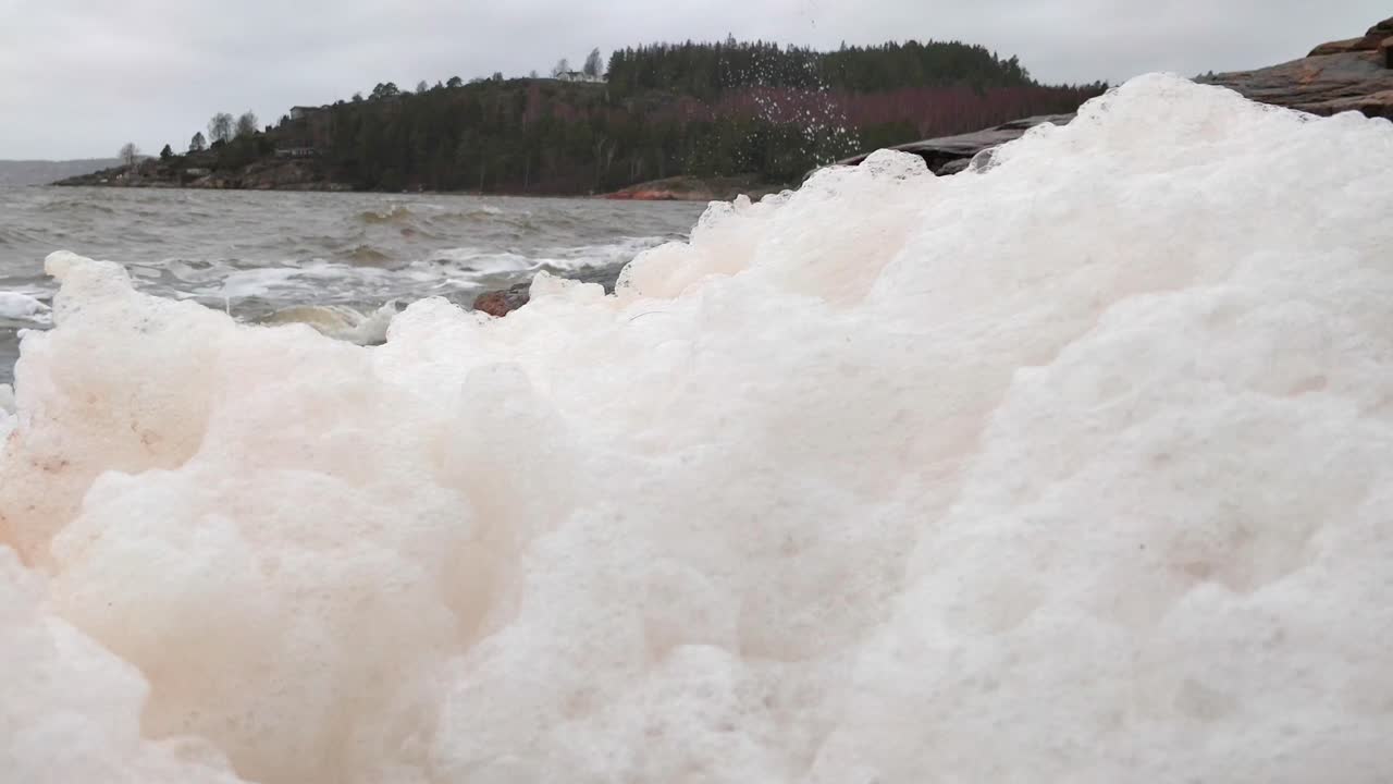 Sea foam sits atop highly agitated water on a grey, rocky coastline