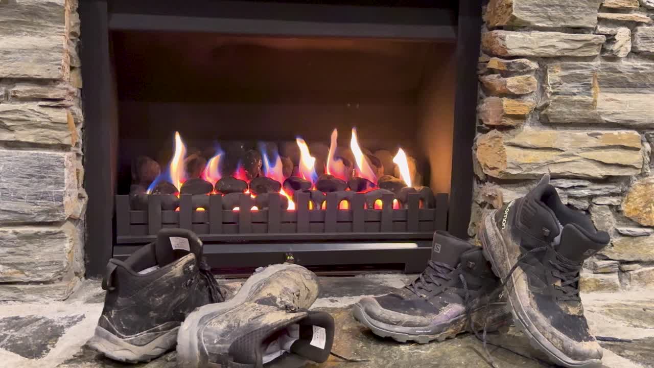 Worn hiking boots drying by warm fireplace, stone surround, steady camera, cozy indoor lighting