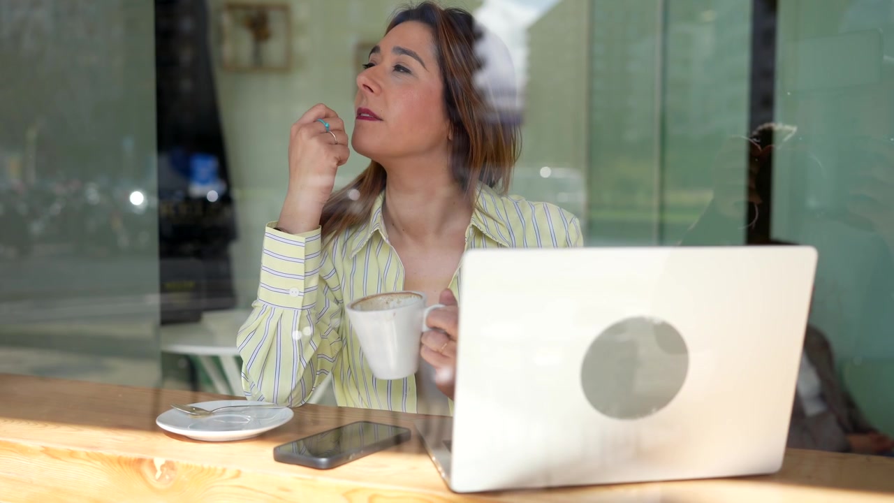 Mujer bebiendo café y mirando por la ventana en una cafetería con portátil