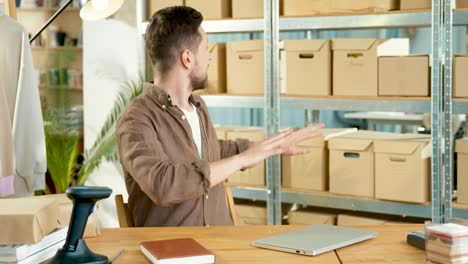 Caucasian-man-sitting-at-desk-at-office-in-clothing-shop-warehouse-speaking-on-video-call-and-looking-at-camera