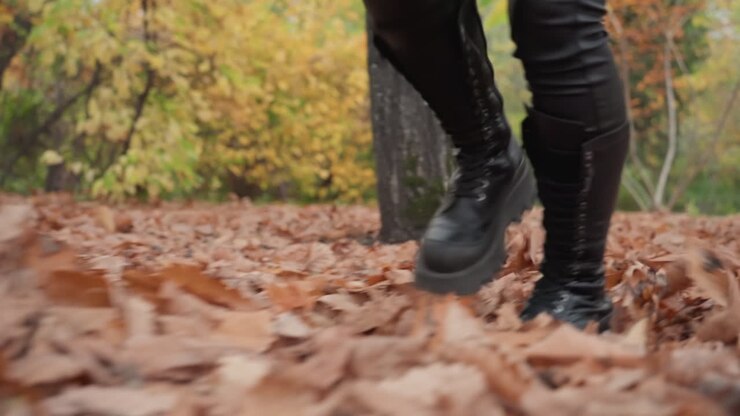Stivali di pelle che camminano in una foresta autunnale, foglie marroni che scricchiolano sotto pesanti stivali stringati, primo piano su pantaloni di pelle e stivali alti, passi al rallentatore, alberi dorati e corteccia ruvida sullo sfondo