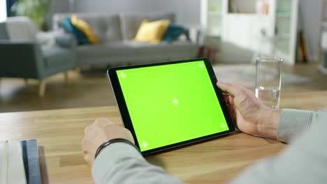 man holding and using hand gestures on green mock-up screen digital tablet computer while sitting at the desk. man buying stuff or browsing through the internet. in the background cozy living room.