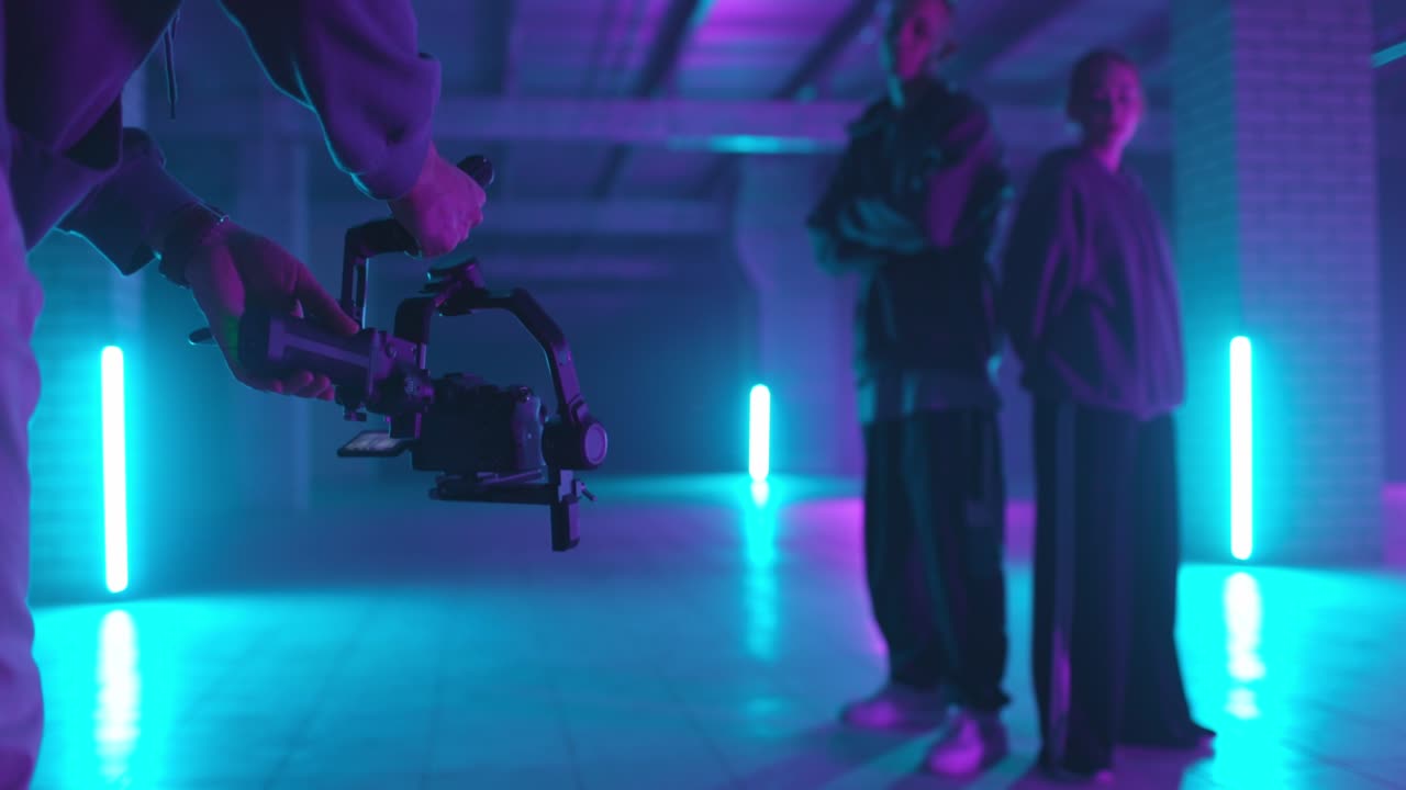 backstage de rodaje de video. camarógrafo filmando bailarines de hip-hop en luz de neón. operador se mueve con estabilizador de imagen en las manos en un estudio oscuro. disparando un clip. vista trasera