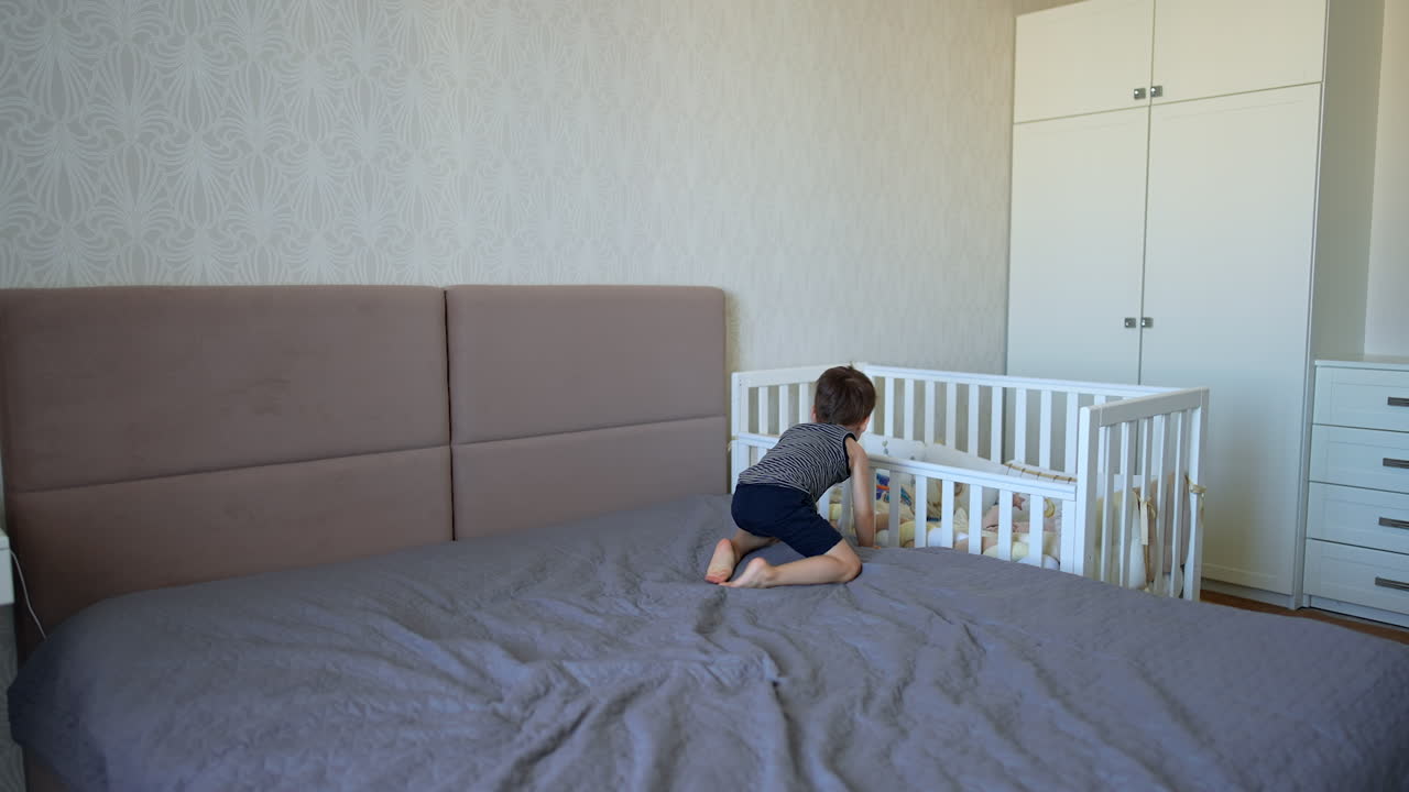 Little baby boy lies on the double bed and tosses his sunglasses. Kid goes to his crib standing beside.