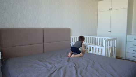little baby boy lies on the double bed and tosses his sunglasses. kid goes to his crib standing beside.