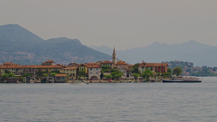 Primo piano di case colorate sull'Isola Borromea con tetti arancioni, una chiesa storica, barche e traghetti di passaggio, con splendide montagne alpine che si affacciano sul Lago Maggiore in Italia