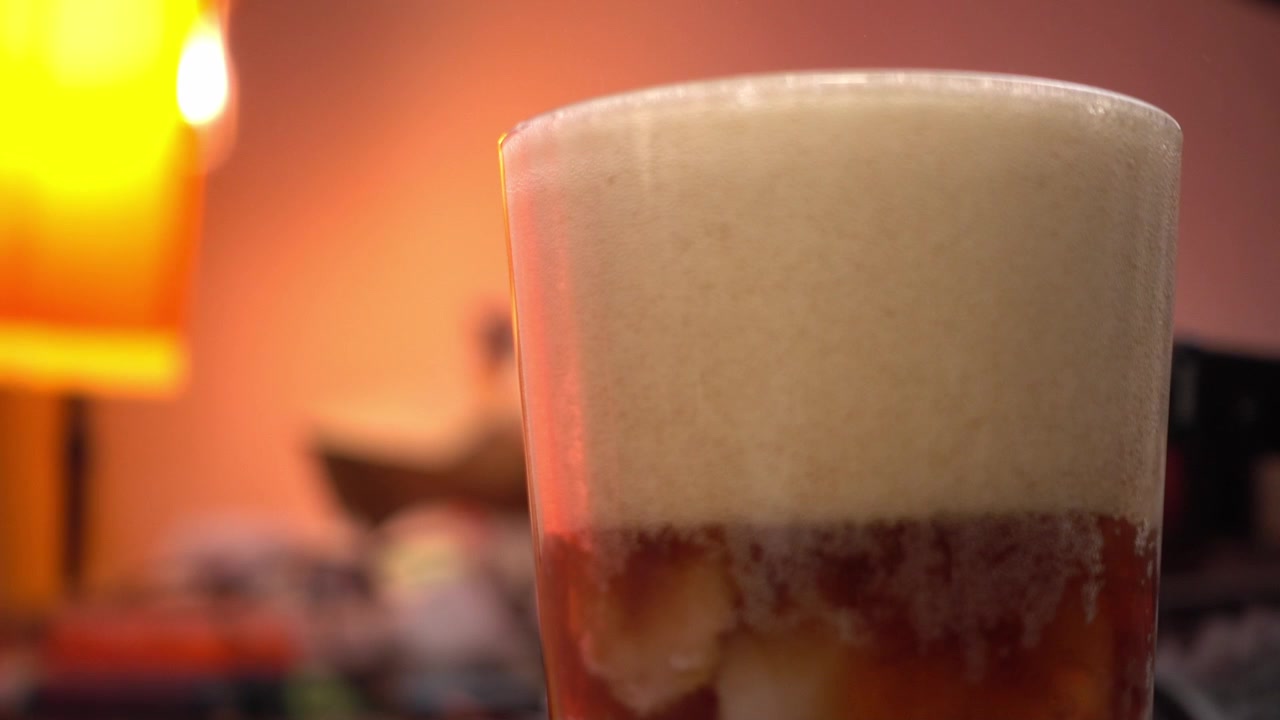 Buz dolu bir bardağa dökülen soda, köpük ve baloncuklar oluştururken, sıcak ve bulanık bir arka planla.