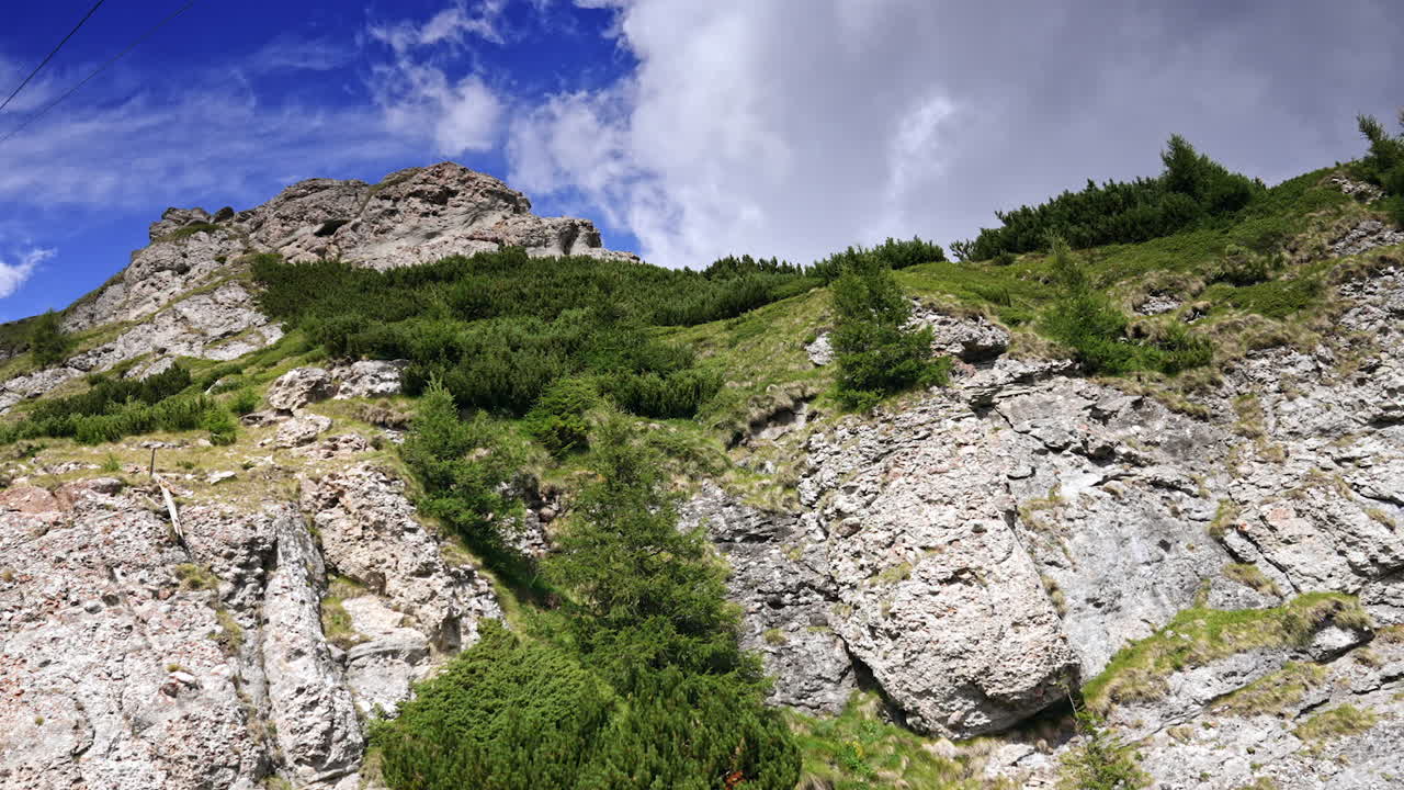 Rocky hill under cloudy sky. Green vegetation clings to a rocky hillside, set against a background of blue sky and white clouds