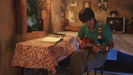 A-young-Asian-male-playing-a-ukulele-while-sitting-on-a-chair-in-a-rustic-room
