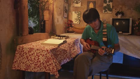 un joven masculino asiático tocando el ukelele mientras está sentado en una silla en una habitación rústica