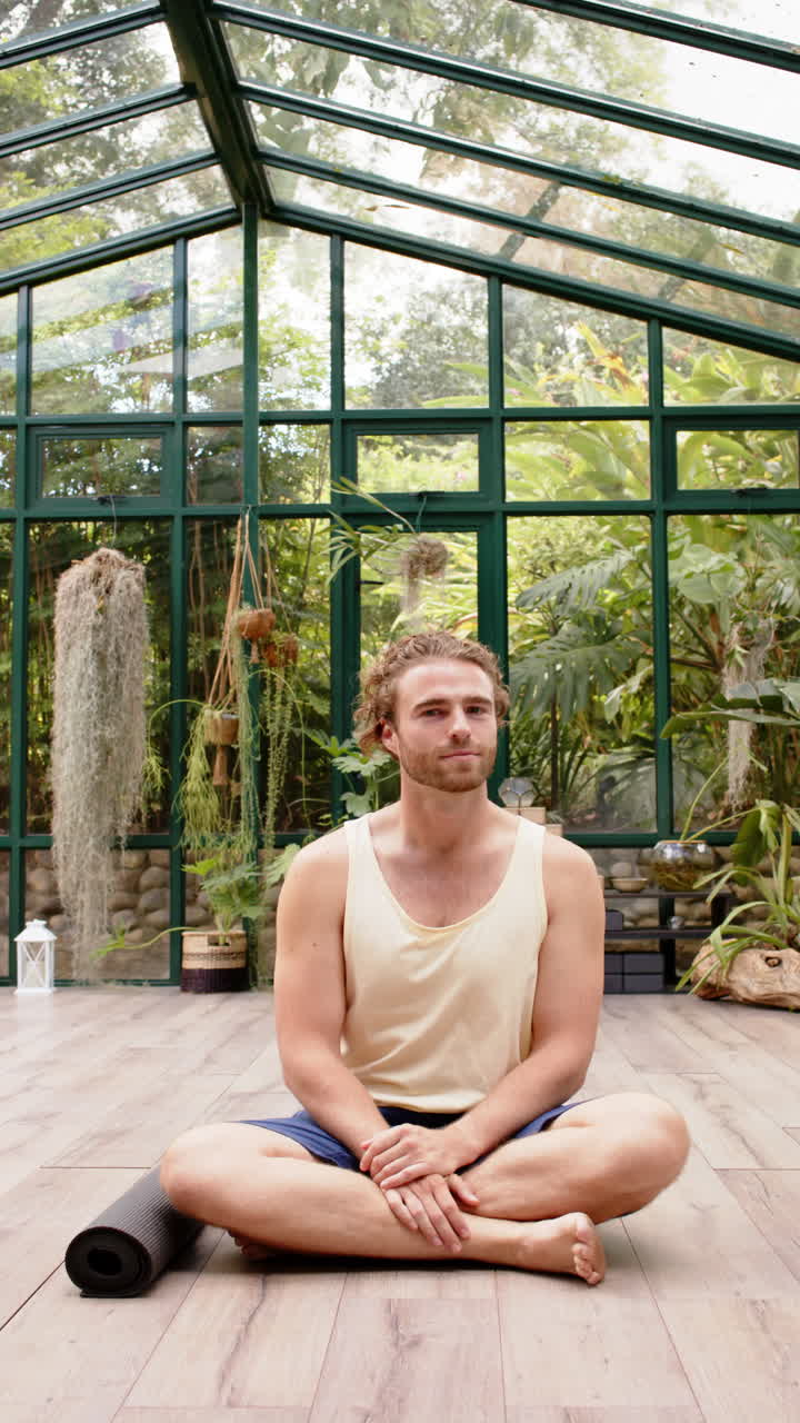 Video verticale: Seduto su un tappetino da yoga, uomo che medita in una casa di vetro circondato da piante