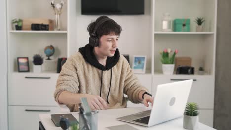 the young man, a freelancer, sits behind his laptop, wearing wireless earphones, in his bright modern room. he stretches because he is tired of working.