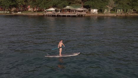 Aerial-drone-bird's-eye-view-of-man-exercising-on-a-sup-paddle-board-in-turquoise-tropical-clear-waters,-with-rocky-coastline-and-backpacker-style-restaurant-in-Koh-Chang-Thailand