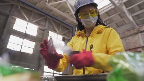 woman-volunteer in yellow and transparent protecting glasses, hard hat and mask sorting used plastic bottles at recycling plant. separate bottles on the line, removing tops and squeeze them. low angle view