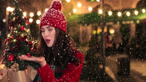 vista de cerca de una mujer caucásica con abrigo rojo sosteniendo un regalo y un árbol de navidad, tratando de atrapar la nieve en la calle en navidad