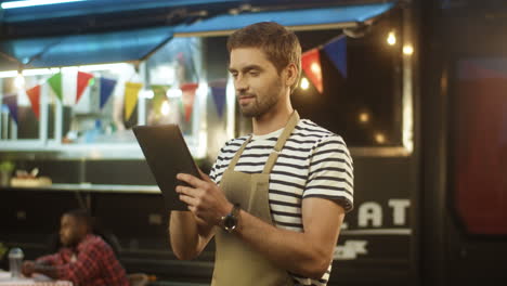 Portrait-Of-Young-Man-Waiter-In-Apron-Smiling-To-Camera-And-Using-Tablet-Device-In-Evening-At-Food-Truck-In-Festive-Park