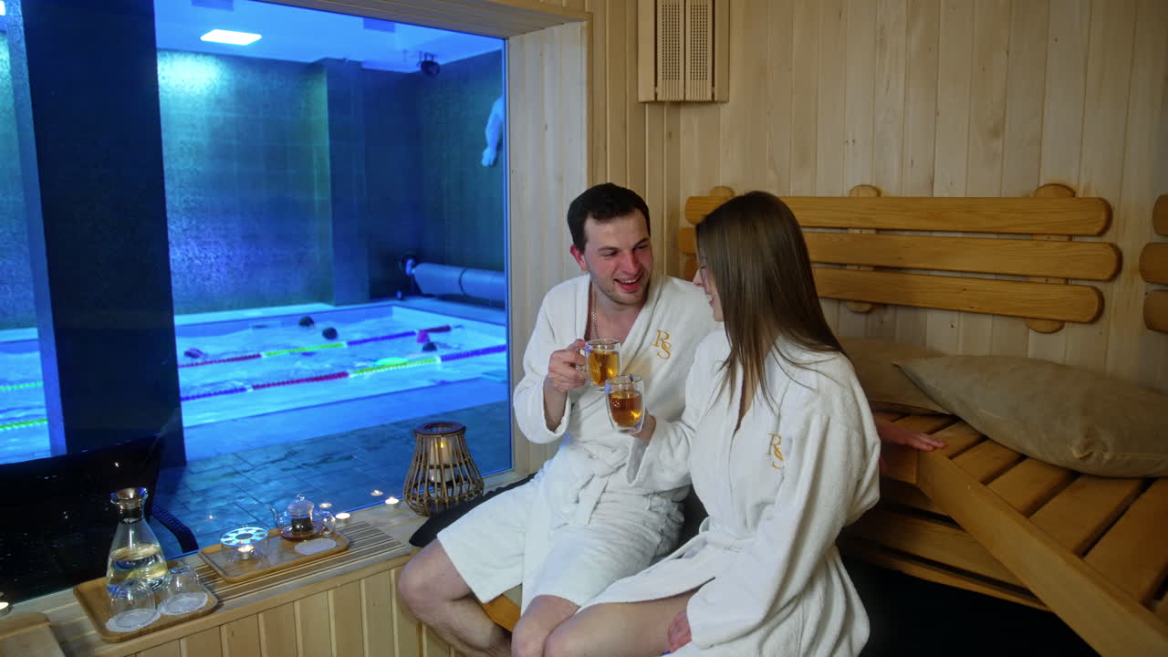 Happy couple wearing the same white soft robes sit in the sauna. Smiling people talk and drink tea from glass double-wall cups. Swimmers in the pool at backdrop.