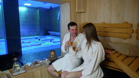 happy couple wearing the same white soft robes sit in the sauna. smiling people talk and drink tea from glass double-wall cups. swimmers in the pool at backdrop.
