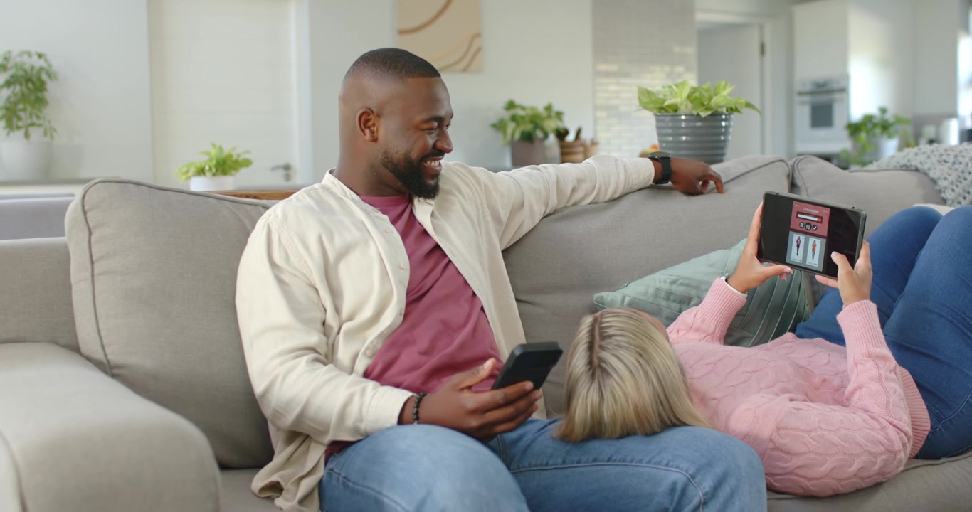 African American couple on sofa, woman raising tablet showing app, man holding smartphone, pointing