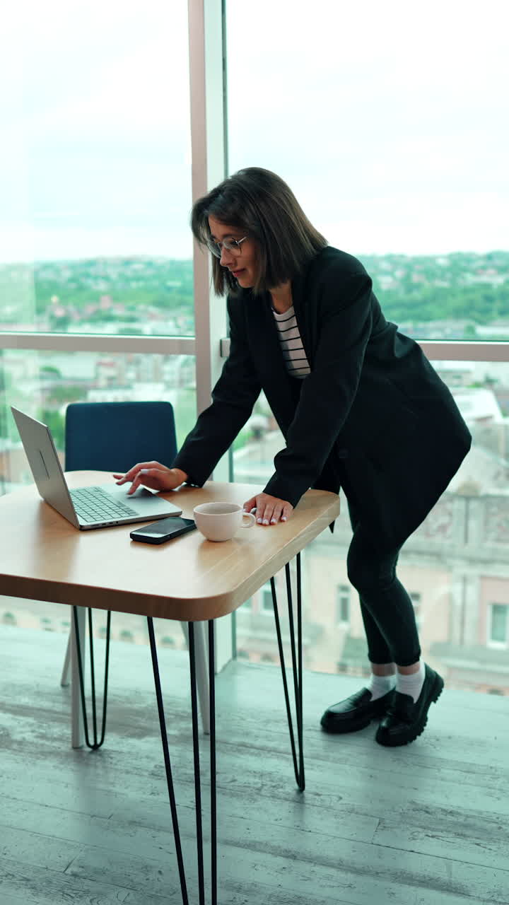 Modern woman stands leaning on the desk using her laptop. Business lady working in the office. Panoramic window at backdrop. Vertical video.