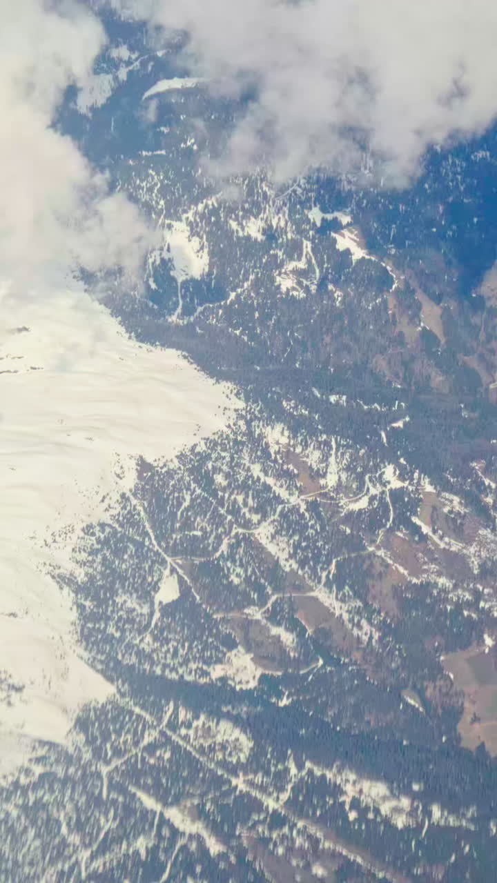 Aerial view of white, fluffy clouds above snowy mountains seen from an airplane window. Vertical