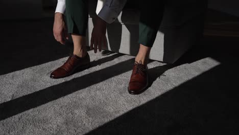 groom tying brown dress shoes, sitting on a bench in sunlight on a carpeted floor