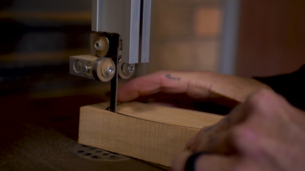 Slow motion close-up of man with hand tattoos carefully cutting wooden climbing hold blocks with electric bandsaw machinery blade tool DIY home garage woodwork shed in Australia carpentry industry