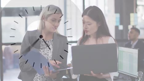 Animation-of-clock-over-two-diverse-businesswomen-using-laptop-talking-in-office