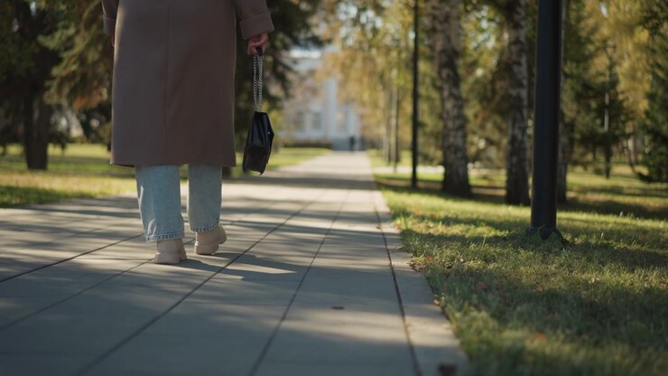 Close up of woman walking down sunlit park pathway holding small black handbag. Woman wears light blue jeans and beige shoes under long brown coat with trees lining path in soft daylight