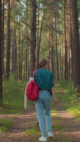 vista de atrás de las damas caminando por un sendero forestal rodeado de árboles altos, la dama de la parte superior verde lleva una mochila roja con un pañuelo drapeado sobre ella, mientras que su compañera en una bufanda azul conduce el camino