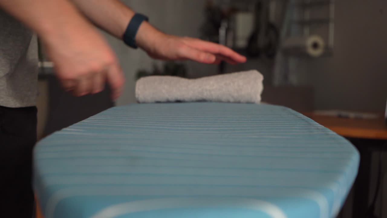 Young Caucasian male folds laundry towels on ironing board, low angle