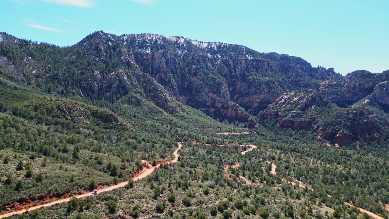 Una vista aerea mostra una strada sterrata rossa che serpeggia tra le montagne boscose dell'Arizona. Un fuoristrada viola percorre il sentiero, con cime innevate sotto un cielo azzurro limpido.