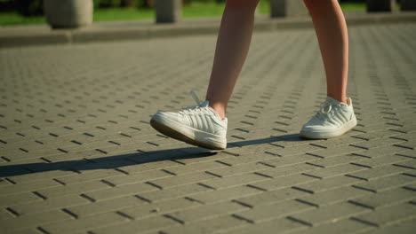 leg view of white lady wearing white canvas shoes walking along interlocked path, shadow cast on ground, with blur background featuring greenery and an unclear object in the distance