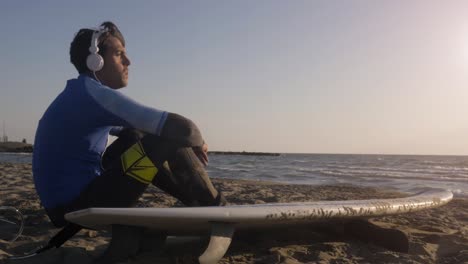 young man surfer in wetsuit sitting with surf board lying down on the sand at the beach listening to music dancing looking at the ocean at sunset