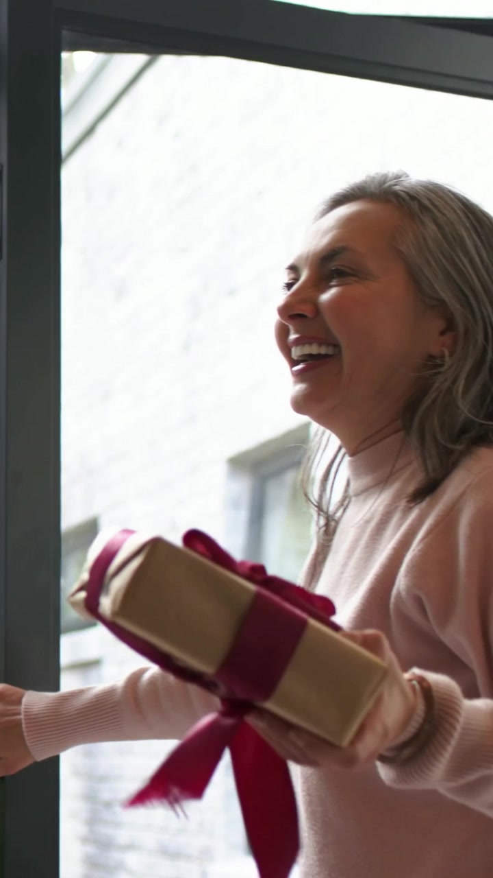 Video verticale: Aprendo la porta, una donna con un maglione rosa porta una scatola impacchettata, visitando la famiglia sulla soglia.