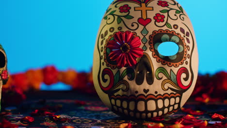 close up of decorated skulls and masks surrounded by flower petals celebrating mexican holiday of dia de muertos or day of the dead against blue background 2