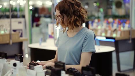 Young-Curly-Woman-In-Glasses-Shopping-For-A-New-Photocamera-In-The-Electronics-Store